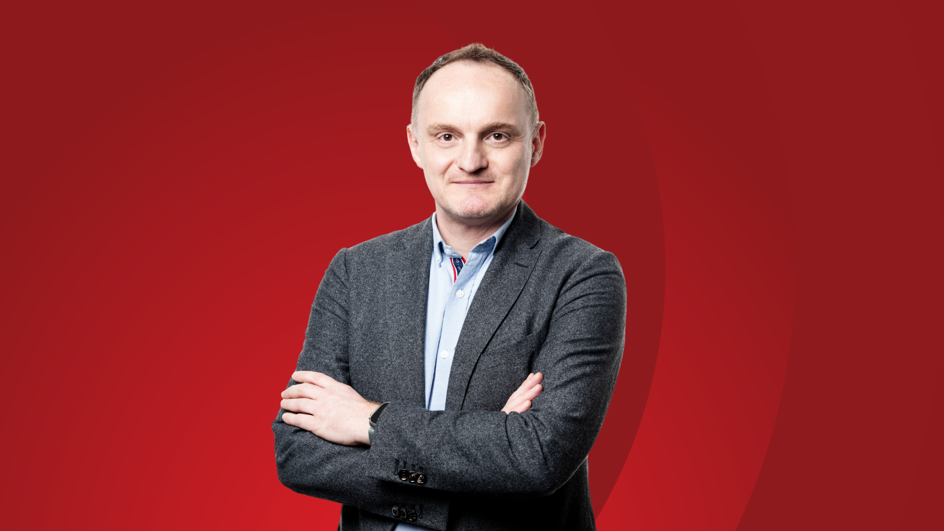The Chief Technology officer, Sylvain Merle stands in a business outfit in portrait mode. The background in a gradient red with part of the logo of BCE.